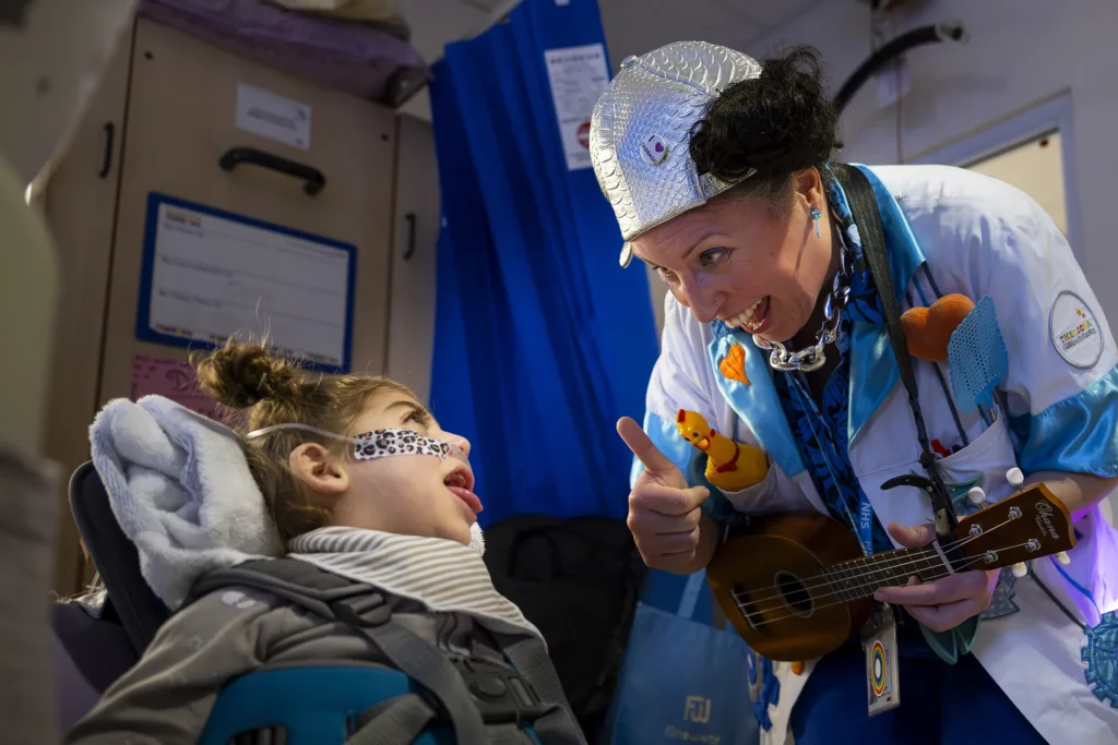 Doctora Sonrisa de la Fundación Theodora toca el ukelele para una niña recostada